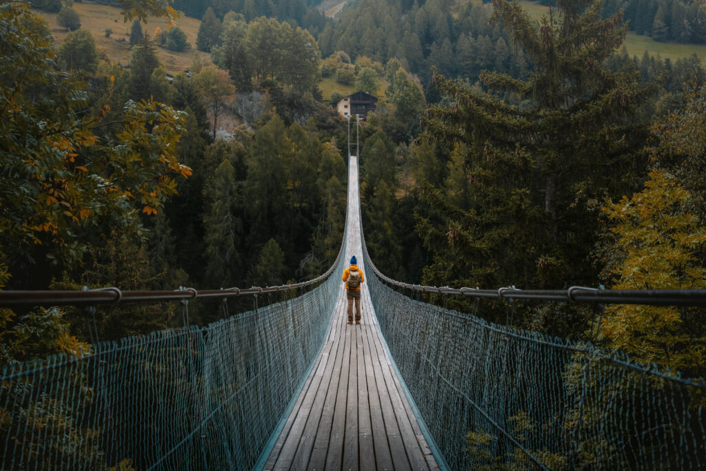 Young traveler man dressed in yellow jacket crosses hiking on an impressive wooden and metal bridge in the village of Goms in the Swiss alps